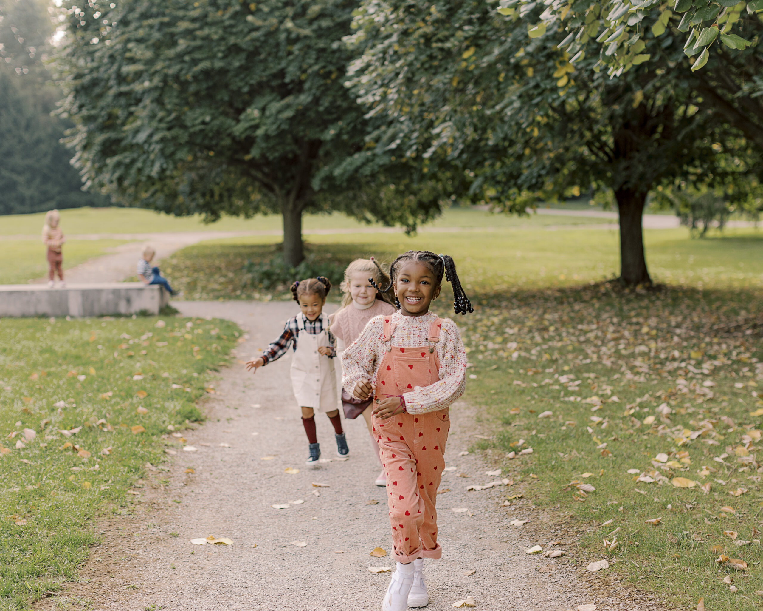 Smiling children running down a leaf-covered park path during a fall mini photo session in Indianapolis, wearing stylish autumn outfits under colorful trees.